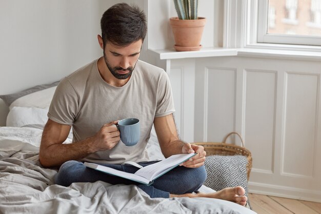 真面目な集中男性は、休日に本を読み、読書に携わり、温かい飲み物を飲み、ベッドに足を組んで座り、カジュアルなTシャツとズボンを着ています。人、知識、教育、レジャー