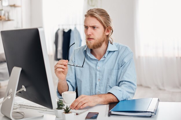 파란색 셔츠에 심각한 집중 잠겨있는 남성 사업가 안경을 손에 보유하고, 컴퓨터에서 작동, 재무 보고서에 대해 생각합니다. 수염 관리자 또는 프리랜서 음료 커피, 아이디어를 생성