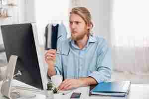Бесплатное фото Серьезный концентрированный задумчивый мужчина бизнесмен в синей рубашке держит очки в руке, работает на компьютере, думает о финансовом отчете. бородатый менеджер или фрилансер пьет кофе, генерирует идеи