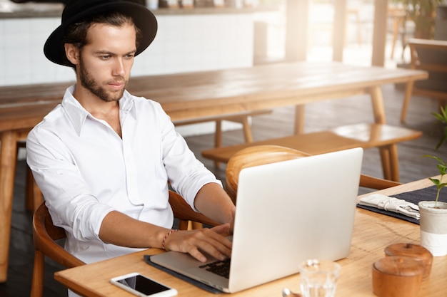 Serious and concentrated young bearded freelancer wearing stylish hat and white shirt using laptop computer for remote work, sitting at cafe table with notebook pc and blank screen mobile phone