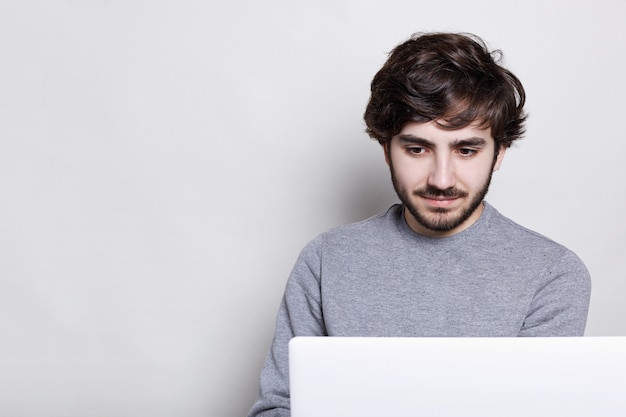 Серьезный и сконцентрированный молодой бородатый парень, носящий повседневный серый свитер, используя портативный компьютер