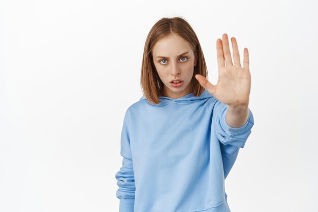 진지하고 자신감 있는 금발 여성이 손을 뻗어 거절 제스처를 금지하고, 아니오라고 말하며, 나쁜 행동을 금지하고, 경고하며, 흰 벽에 기대어 서 있습니다.