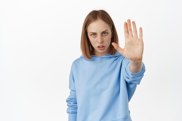 Serious and confident blond woman stretch out her hand, showing prohibit rejection gesture, say no, forbid bad action, warn you, standing against white wall