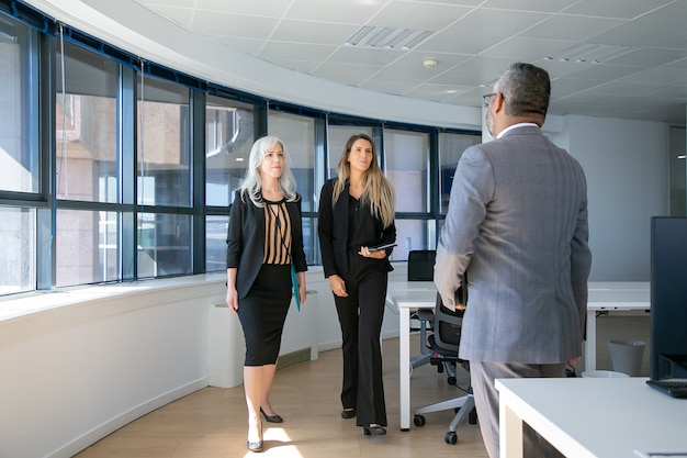 Serious confident businesswomen walking to man in suit in office interior. Full length, back view. Business meeting concept