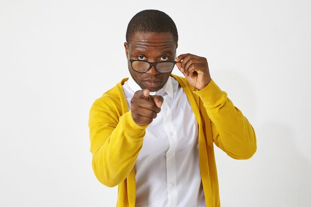 Serious confident dark skinned male teacher in eyewear pointing index finger , having strict look, warning his students, standing isolated at white  wall with copyspace for your text