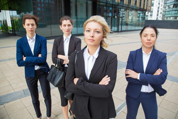 深刻な自信を持って女性のプロのチームが一緒に事務所ビルの近くのグループリーダーと一緒に立って、ポーズをとって、カメラ目線します。正面図。ビジネスウーマングループの肖像画のコンセプト