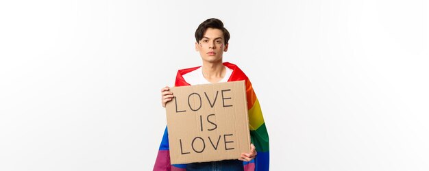 Free photo serious and confident gay man wearing rainbow lgbt flag holding sign for pride parade standing over