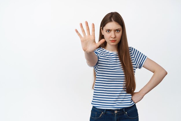 Serious and confident girl stretch out hand to stop, showing prohibition, warning gesture, taboo sign, standing on white