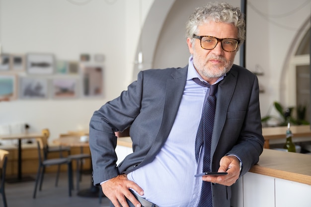 Serious confident mature businessman wearing glasses and suit, using smartphone, standing in co-working space