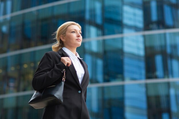 Serious confident middle aged business lady wearing office suit, holding bag, walking past glass office building. Low angle, copy space. Businesswoman in city concept