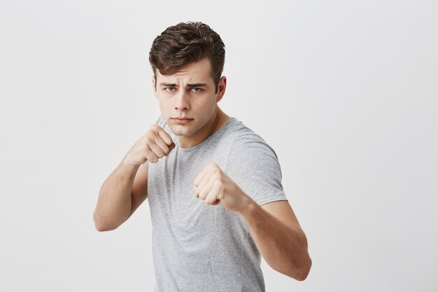 Serious confident muscular young sportsman frowns face in displeasure, shows clenched fists, demonstrates strength and irritation, ready to defend himself. Power and strenght concept.