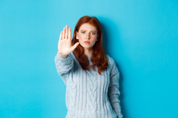 Serious and confident redhead girl telling to stop, saying no, showing extended palm to prohibit action, standing over blue background