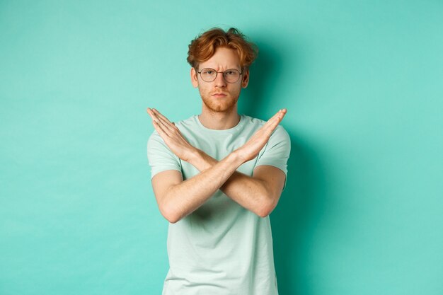 Serious and confident redhead man in t-shirt and glasses saying no, showing cross gesture to stop you, refucing or declining something, standing over turquoise background.