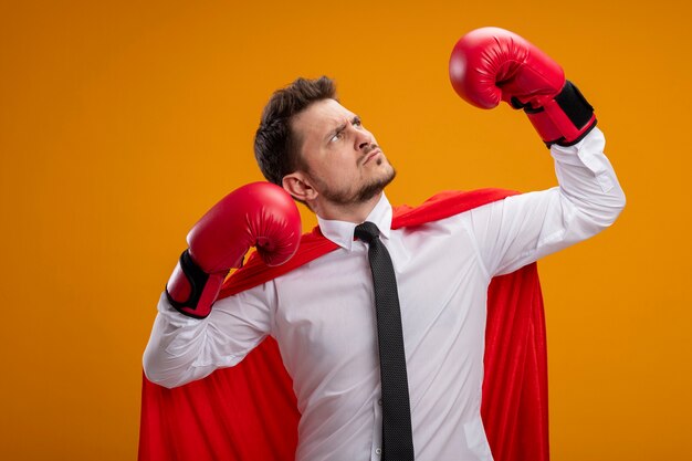 Serious confident super hero businessman in red cape and in boxing gloves raising hands showing strength and courage standing over orange background