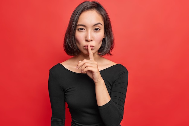 Серьезная уверенная в себе молодая азиатская женщина делает жест молчания, держит указательный палец над губами, рассказывает секретную или конфиденциальную информацию, носит черное платье с длинными рукавами, изолированное над ярко-красной стеной.