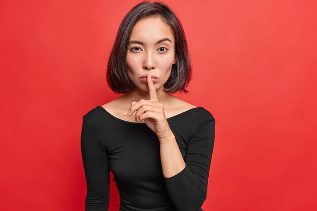 무료 사진 진지하고 자신감 있는 젊은 아시아 여성은 입술에 검지 손가락을 대고 조용히 몸짓을 하며 비밀 정보를 말하거나 밝은 빨간색 벽에 격리된 검은색 긴 소매 드레스를 입습니다.