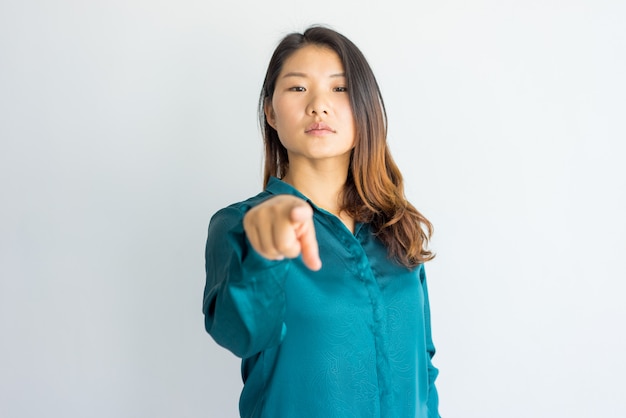 Serious confident young Asian woman pointing at you.