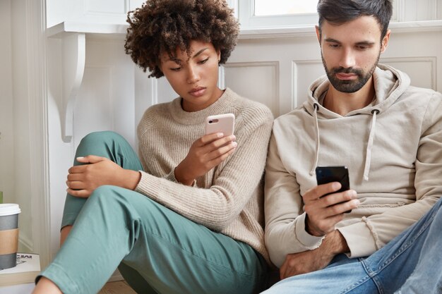 Serious couple use modern technologies, wireless connection at home, send text messages on smart phones