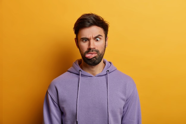 Serious crazy bearded man crosses eyes and sticks out tongue, has funny grimace, feels bored, raises eyebrows, wears casual hoodie, isolated on yellow wall. human face expressions concept