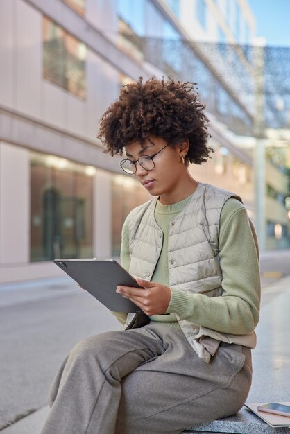 Serious curly female student studies online uses modern teblet wears spectacles casual clothes browses internet poses outside against blurred background Freelancer thinks about strategy of project