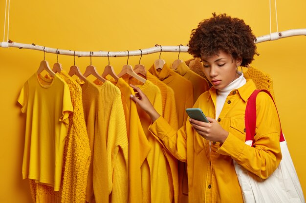 Serious curly haired female buyer carries bag, picks up yellow clothes, concentrated in smartphone, poses near rack of stylish clothing, makes purchase