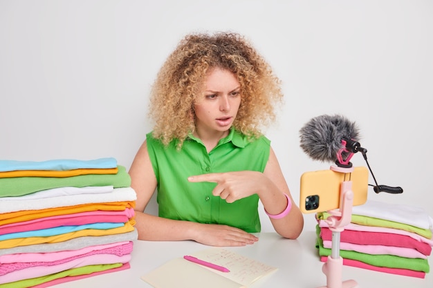 Serious curly haired female vlogger points at pile of multicolored folded laundry dressed in green t shirt talks about softener for your washing sits at table gives advice how to choose detergent