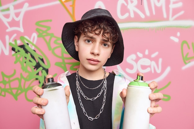 Free photo serious curly haired hipster wears hat shirt metal chains around neck holds two aerosol sprays draws graffiti wall being street artist has creative hobby activity