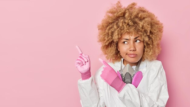 Серьезная кудрявая женщина-ученый носит перчатки защитного костюма и респиратор на шее рассказывает о токсичных отходах и загрязнении на месте экологического бедствия указывает на пустое пространство у розовой стены