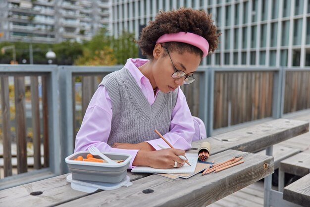 Serious curly haired young woman makes notes wears round spectacles neat clothes has delicious snack works on creative task or project poses outdoors going to have healthy lunch makes planning