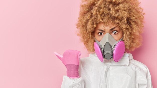 Serious curly haired young woman points away on blank space wears protective suit and respirator shows place for your advertisement or promotion warns you about radiation or biochemical hazard