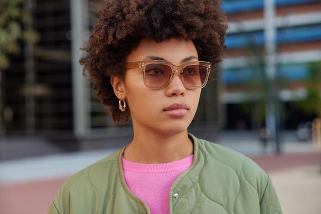 Serious curly haired young woman wears sunglasses and jacket has promenade in urban setting concentrated into distance poses outdoors against blurred background. Human fashion style concept.