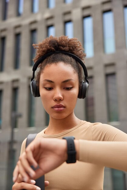 Serious curly sportswoman checks results of workout on smartwatch listens music in wireless headphones dressed in activewear