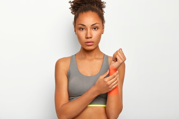 Serious curly young woman with dark skin massages her arthritic hand and wrist, injured arm after sport training, touches red spot, indicates location of pain, wears grey sport bra, poses indoor