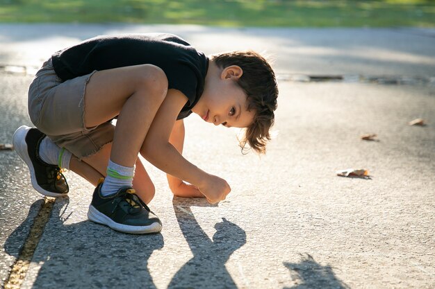 Serious cute boy drawing on playground surface with colorful pieces of chalks. Side view. Childhood and creativity concept