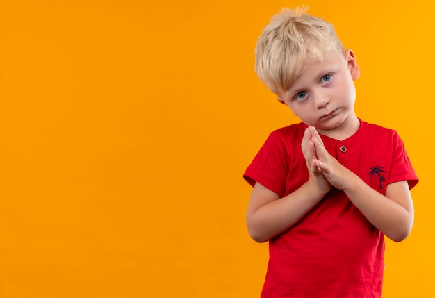 A serious cute little boy with blonde hair and blue eyes wearing red t-shirt looking while holding hand together on a yellow wall