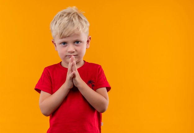 Free photo a serious cute little boy with blonde hair and blue eyes wearing red t-shirt looking on a yellow wall