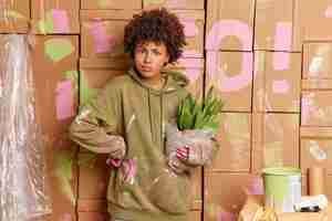 Free photo serious dark skinned curly woman busy renovating house wears casual sweatshirt dirty with paint painting walls in apartment plans home repair holds pot of green cactus. house repair concept.