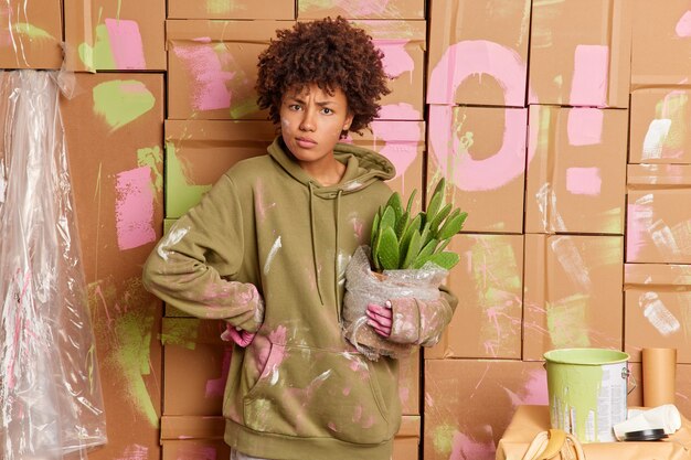 Serious dark skinned curly woman busy renovating house wears casual sweatshirt dirty with paint painting walls in apartment plans home repair holds pot of green cactus. House repair concept.