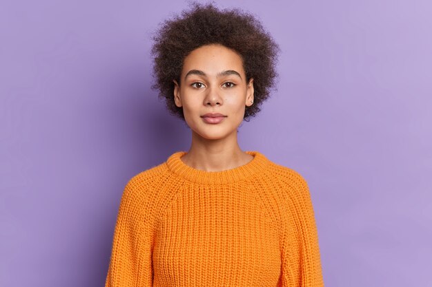 serious dark skinned female teenager with curly bushy hair looks confidently has natural beauty calm expression dressed in knitted sweater.