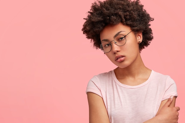 Grave femmina dalla pelle scura con capelli croccanti, vestita con una maglietta casual, ha la pelle scura, posa contro il muro rosa con copyspace