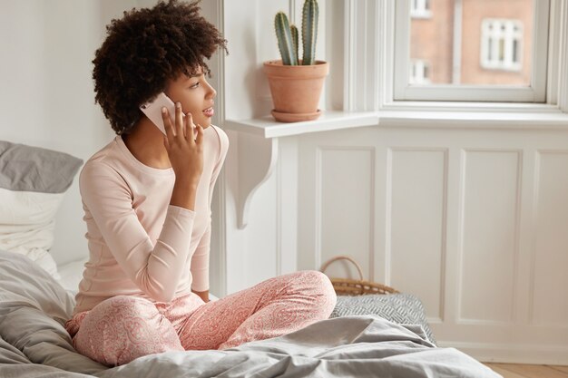 Serious dark skinned girl makes telephone call, satisfied with tariffs and internet connection at home, wears casual nightwear, looks aside