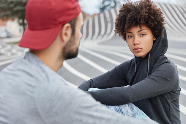 Free photo serious dark skinned lady in hoodie, sits opposite her best male friend