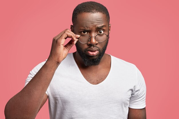 Serious dark skinned male professor looks in bewilderment through spectacles