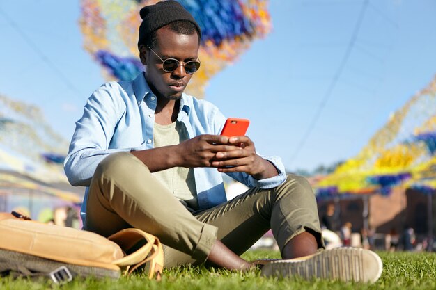 트렌디 한 셔츠와 바지에 심각한 뭇 젊은 남자, 녹색 잔디에서 휴식, 손에 빨간 스마트 폰을 들고, 인터넷에서 무언가를 읽거나 메시지를 입력. 편안한 검은 유행 남성