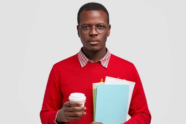 Серьезный смуглый молодой человек с уверенным выражением лица, готовый к учебе, держит учебники и берет кофе на вынос, позирует над белым пространством