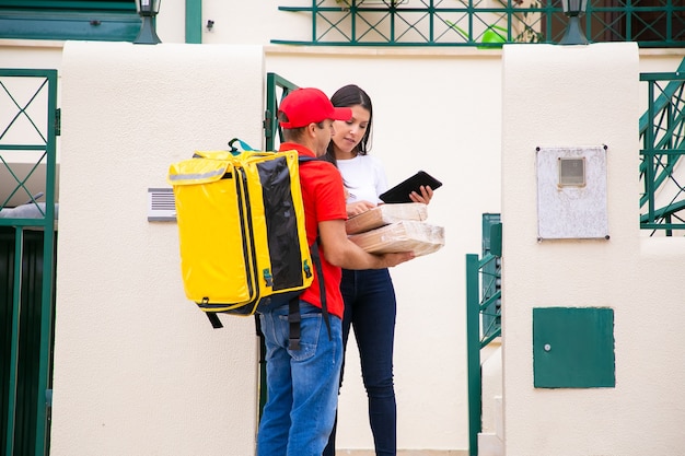 小包を持っている真面目な配達員と注文をチェックしている女性。赤いキャップと黄色の保温バッグが付いたシャツのコンテンツ宅配便で、徒歩でエクスプレスオーダーを提供します。配送サービスとポストコンセプト