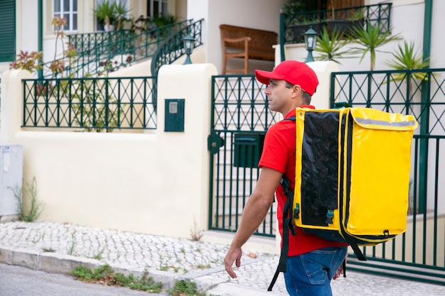 Serious deliveryman looking for address and carrying yellow thermal bag. Attractive courier in red shirt walking along street and delivering order. Food delivery service and online shopping concept
