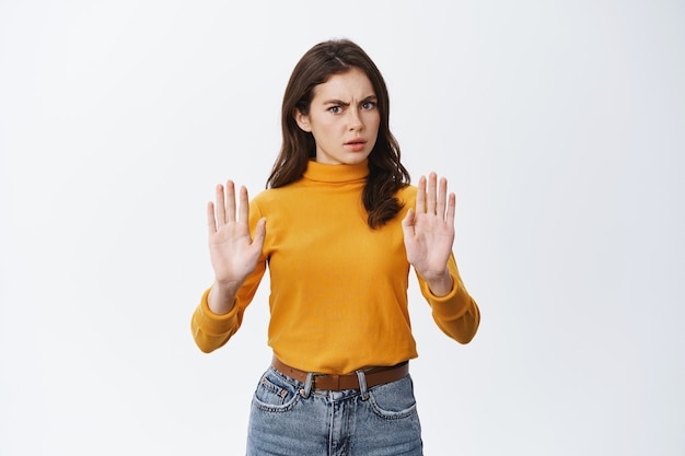 La donna seria e determinata dice di no, mostrando il gesto di proibire e proibire, chiedendo di calmarsi o rifiutare qualcosa di brutto, accigliato, in piedi sul muro bianco