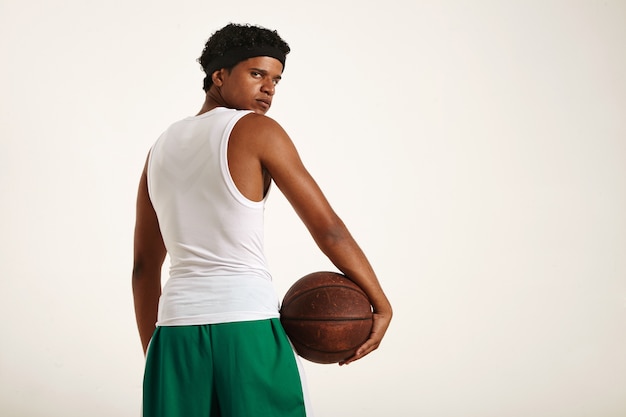 Serious determined young African American basketball player in white and green uniform with a short afro holding an old brown basketball to his hip and looking back