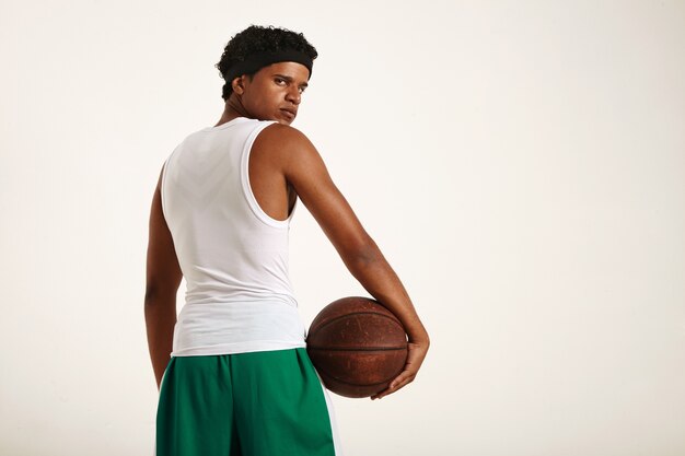 Серьезный и целеустремленный молодой афроамериканский баскетболист в бело-зеленой форме с коротким афро, держащим старый коричневый баскетбольный мяч у бедра и оглядывающимся назад
